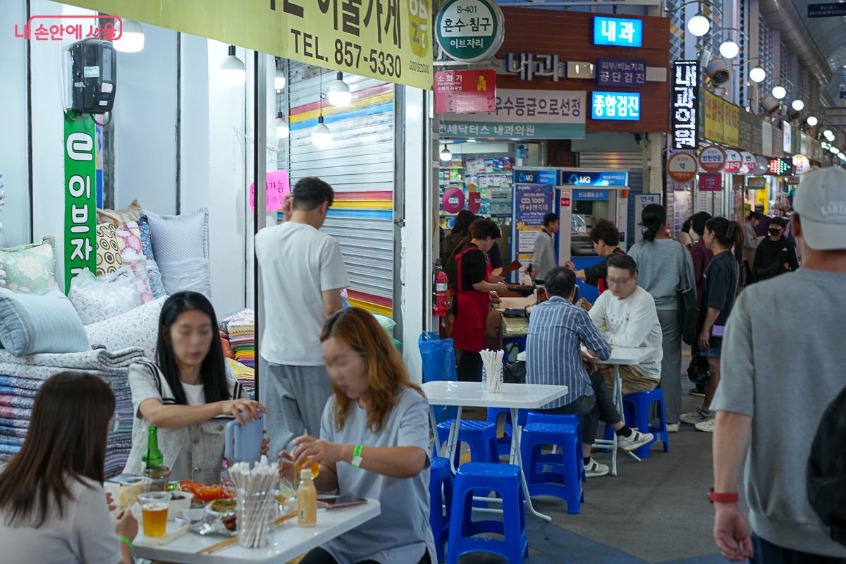 저녁 불빛이 켜진 아케이드 아래, 파란 간이 의자에 둘러앉아 음료를 마시며 이야기를 나누는 방문객들