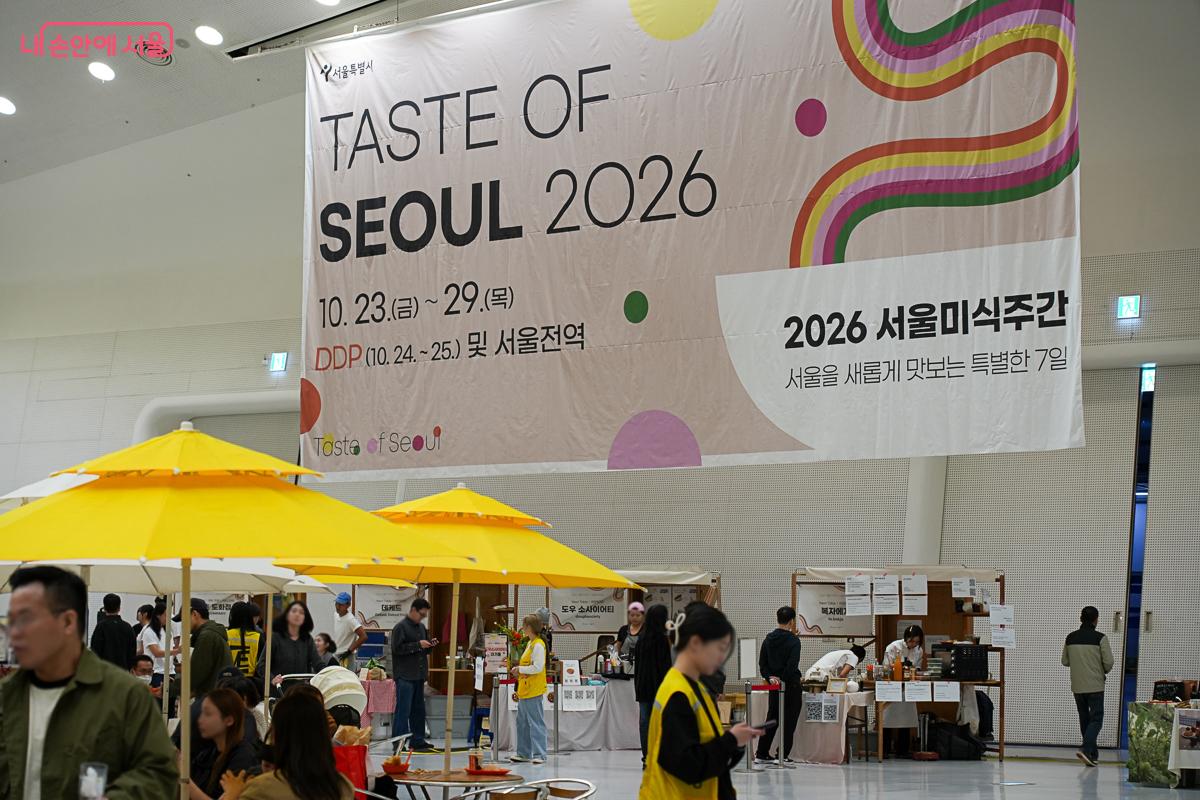 DDP 실내 공간 천장에 'TASTE OF SEOUL 2026 / 2026 서울미식주간' 대형 현수막이 걸려 있고, 아래로 노란 파라솔 테이블에서 시민들이 식사하고 있다.